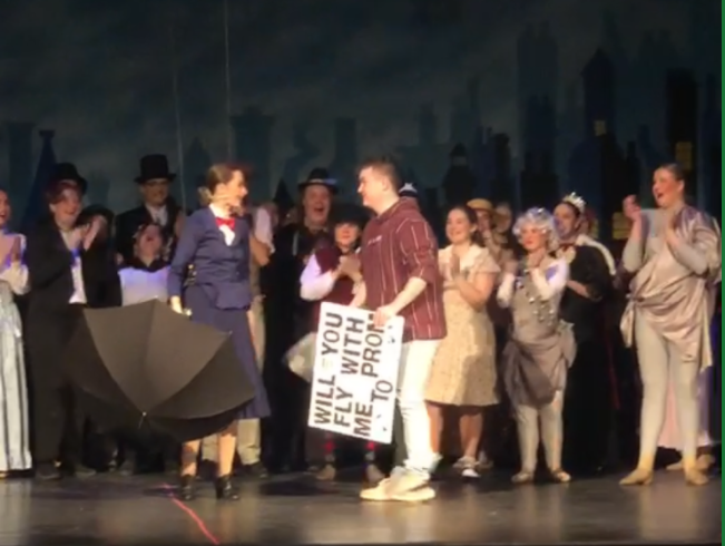 Alana+Weirbach+and+Ian+Meschester+on+stage+during+their+promposal.+Meschester+holds+a+bouquet+of+flowers+and+a+sign+saying+will+you+fly+with+me+to+prom%3F